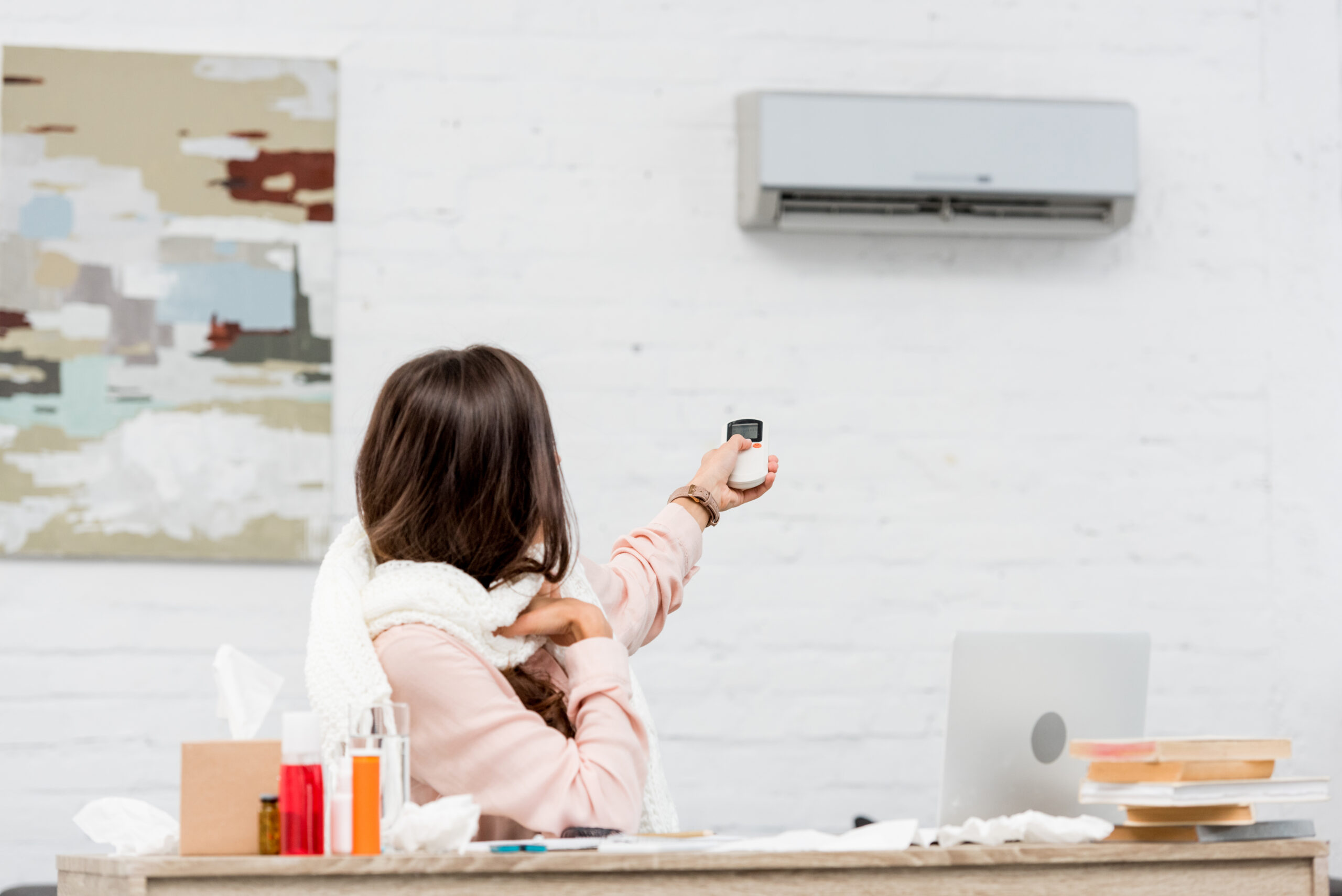 sick young woman sitting at workplace and pointing at air conditioner with remote control